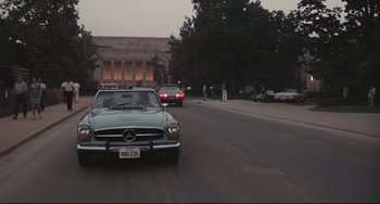 Movie still from “Breaking Away” (1979), directed by Peter Yates – A car driving down a street next to a building; Extreme Wide shot, Over the shoulder angle