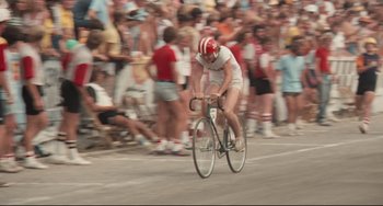 Movie still from “Breaking Away” (1979), directed by Peter Yates – A man riding a bike down a street with a crowd of people behind him; Wide shot, Low angle