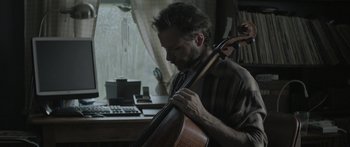 Movie still from “Breathe In” (2013), directed by Drake Doremus – A man holding a cello in front of a computer desk; Medium shot, Over the shoulder angle