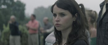 Movie still from “Breathe In” (2013), directed by Drake Doremus – A young woman is looking at the camera while standing in front of a group of people; Close Up shot, Over the shoulder angle