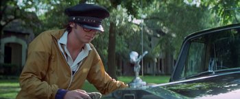 Movie still from “Brewster McCloud” (1970), directed by Robert Altman – A man in a hat and glasses is standing next to an antique car; Medium shot, Low angle