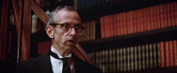 Movie still from “Brewster McCloud” (1970), directed by Robert Altman – An older man wearing a suit and tie in front of a book shelf; Close Up shot, Over the shoulder angle