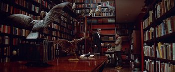 Movie still from “Brewster McCloud” (1970), directed by Robert Altman – A man in a suit standing in front of an owl statue in a room full of bookshelves; Wide shot, Low angle