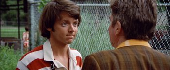 Movie still from “Brewster McCloud” (1970), directed by Robert Altman – A young man and an older man are standing next to each other in front of a chain link fence; Close Up shot, Over the shoulder angle