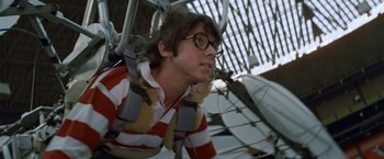 Movie still from “Brewster McCloud” (1970), directed by Robert Altman – A young man wearing glasses and a striped shirt hanging from a parachute; Medium shot, Low angle