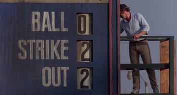 Movie still from “Brewster's Millions” (1985), directed by Walter Hill – A man leaning up against the side of a baseball field; Wide shot, Low angle