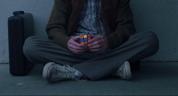 Movie still from “Brick” (2005), directed by Rian Johnson – A man sitting on the ground holding a rubiks cube; Medium shot, Overhead angle