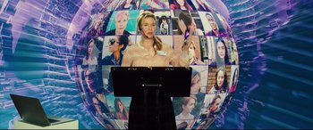 Movie still from “Bridget Jones's Baby” (2016), directed by Sharon Maguire – A woman standing at a podium in front of a wall of pictures; Medium shot, Overhead angle