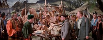 Movie still from “Brigadoon” (1954), directed by Vincente Minnelli – A group of people standing next to a table with food on top of it; Medium shot, High angle