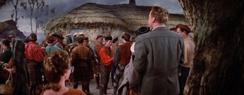 Movie still from “Brigadoon” (1954), directed by Vincente Minnelli – A group of people standing in front of a thatched roof house; Medium shot, Over the shoulder angle