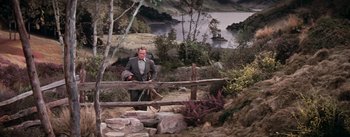 Movie still from “Brigadoon” (1954), directed by Vincente Minnelli – A man in a gray suit is standing by a wooden fence; Wide shot, High angle