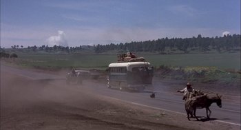 Movie still from “Bring Me the Head of Alfredo Garcia” (1974), directed by Sam Peckinpah – A bus is driving down a road with a car on top of it; Extreme Wide shot, High angle