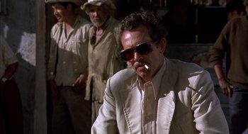 Movie still from “Bring Me the Head of Alfredo Garcia” (1974), directed by Sam Peckinpah – A man in a white suit smoking a cigarette in front of a group of men; Medium shot, Over the shoulder angle