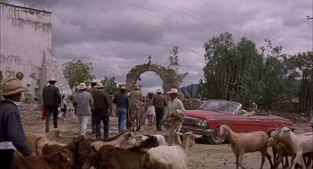 Movie still from “Bring Me the Head of Alfredo Garcia” (1974), directed by Sam Peckinpah – A group of people standing next to a red car and goats; Extreme Wide shot, High angle