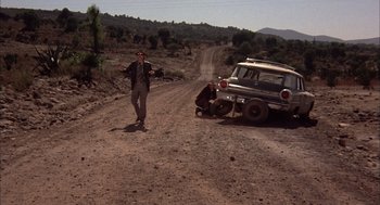Movie still from “Bring Me the Head of Alfredo Garcia” (1974), directed by Sam Peckinpah – A man standing next to a car on the side of a dirt road; Wide shot, Low angle