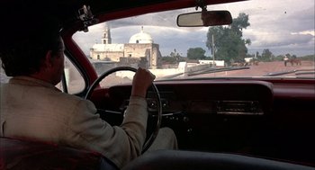 Movie still from “Bring Me the Head of Alfredo Garcia” (1974), directed by Sam Peckinpah – A man driving a red car on a street; Wide shot, Over the shoulder angle