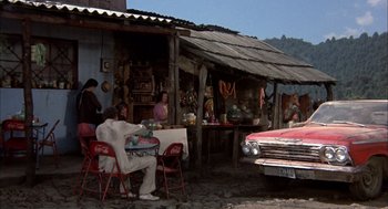 Movie still from “Bring Me the Head of Alfredo Garcia” (1974), directed by Sam Peckinpah – A group of people sitting at a table in front of an old car; Wide shot, High angle