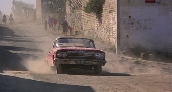 Movie still from “Bring Me the Head of Alfredo Garcia” (1974), directed by Sam Peckinpah – An old car driving down a dirt road with people walking around; Wide shot, High angle