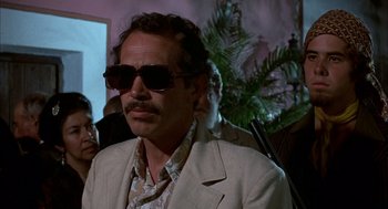 Movie still from “Bring Me the Head of Alfredo Garcia” (1974), directed by Sam Peckinpah – A man in a suit and sunglasses is sitting in front of a group of people; Close Up shot, Low angle