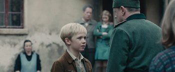 Movie still from “The Book Thief” (2013), directed by Brian Percival – A young boy with blonde hair standing in front of a group of people; Medium shot, Over the shoulder angle