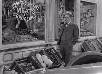 Movie still from “Bringing Up Baby” (1938), directed by Howard Hawks – A man standing in front of a store with produce in front of it; Medium shot, Over the shoulder angle