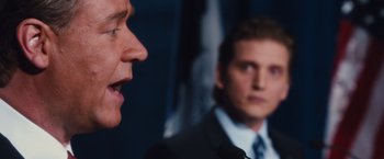 Movie still from “Broken City” (2013), directed by Allen Hughes – A man in a suit and tie talking into a microphone; Close Up shot, Over the shoulder angle