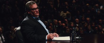 Movie still from “Broken City” (2013), directed by Allen Hughes – A man sitting at a table in front of an audience; Medium shot, Low angle