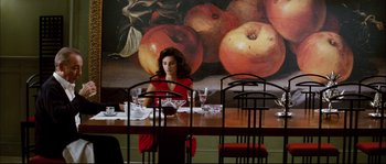 Movie still from “Broken Embraces” (2009), directed by Pedro Almodóvar – A woman sitting at a table with a glass of wine in front of a painting of apples; Wide shot, High angle
