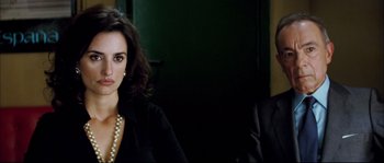 Movie still from “Broken Embraces” (2009), directed by Pedro Almodóvar – A woman sitting next to a man wearing a suit and pearl necklace; Close Up shot, Over the shoulder angle