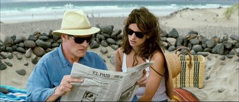 Movie still from “Broken Embraces” (2009), directed by Pedro Almodóvar – A man and a woman sitting on the beach looking at a newspaper; Medium shot, High angle