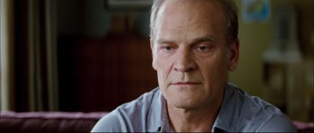 Movie still from “Broken Embraces” (2009), directed by Pedro Almodóvar – An older man with a goatee looking at the camera; Close Up shot, Over the shoulder angle