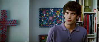 Movie still from “Broken Embraces” (2009), directed by Pedro Almodóvar – A man in a purple polo shirt in front of a floral painting; Close Up shot, Over the shoulder angle