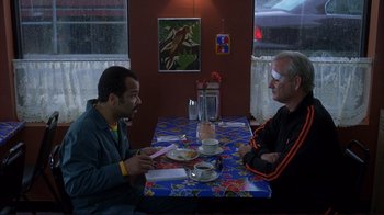 Movie still from “Broken Flowers” (2005), directed by Jim Jarmusch – Two men are sitting at a table in front of a painting; Medium shot, Over the shoulder angle