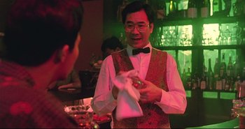 Movie still from “Bullet in the Head” (1990), directed by John Woo – A man in a red vest and bow tie holding a white towel; Medium shot, Over the shoulder angle