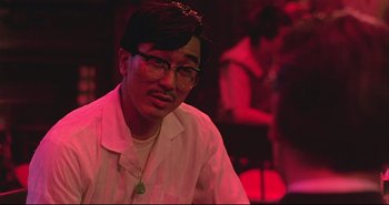 Movie still from “Bullet in the Head” (1990), directed by John Woo – A man with glasses is sitting in front of a crowd; Close Up shot, Over the shoulder angle