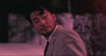 Movie still from “Bullet in the Head” (1990), directed by John Woo – A man in a suit and tie standing in front of a building; Close Up shot, Low angle