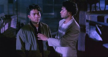 Movie still from “Bullet in the Head” (1990), directed by John Woo – Two men are standing in the dark and one of them has his hand up to his chest; Medium shot, Over the shoulder angle
