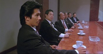 Movie still from “Bullet in the Head” (1990), directed by John Woo – A group of men sitting at a long wooden table; Medium shot, Over the shoulder angle