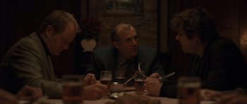 Movie still from “Bullhead” (2011), directed by Michaël R. Roskam – A group of people sitting around a table with drinks; Medium shot, Over the shoulder angle