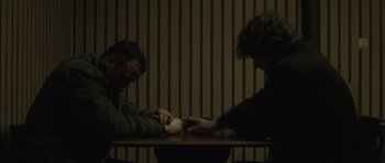 Movie still from “Bullhead” (2011), directed by Michaël R. Roskam – Two people sitting at a wooden table in a dark room; Medium shot, High angle