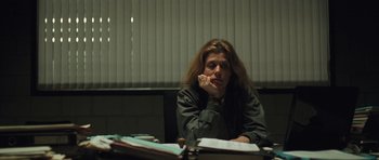 Movie still from “Bullhead” (2011), directed by Michaël R. Roskam – A woman sitting at a desk with her head resting on her hand; Medium shot, Over the shoulder angle