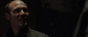 Movie still from “Bullhead” (2011), directed by Michaël R. Roskam – A man in a dark room smiling at the camera; Close Up shot, Over the shoulder angle