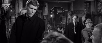 Movie still from “Bunny Lake Is Missing” (1965), directed by Otto Preminger – A man standing in front of a crowd of people in front of a building; Medium shot, Over the shoulder angle