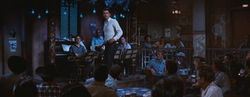 Movie still from “Bus Stop” (1956), directed by Joshua Logan – A group of people sitting in a room with a man standing in front of them; Wide shot, Over the shoulder angle