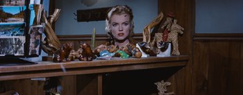 Movie still from “Bus Stop” (1956), directed by Joshua Logan – A woman sitting in front of a wooden table with many figurines on top of it; Close Up shot, High angle