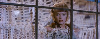 Movie still from “Bus Stop” (1956), directed by Joshua Logan – A woman standing in front of a window with lace curtains; Medium shot, Low angle