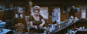 Movie still from “Bus Stop” (1956), directed by Joshua Logan – A woman in a green dress is cooking in a kitchen; Medium shot, Over the shoulder angle