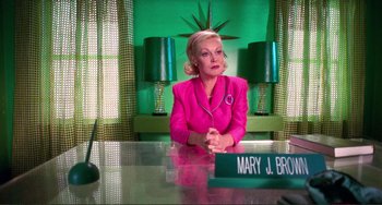 An older woman sitting at a desk in front of a green wall