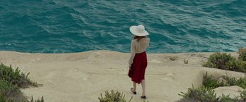 Movie still from “By the Sea” (2015), directed by Angelina Jolie – A woman in a white hat and a red skirt standing on a cliff by the water; Wide shot, High angle