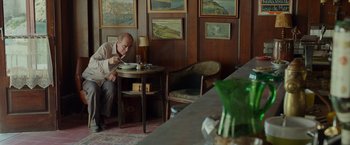 Movie still from “By the Sea” (2015), directed by Angelina Jolie – An older man sitting at a table with a plate in front of him; Medium shot, Over the shoulder angle